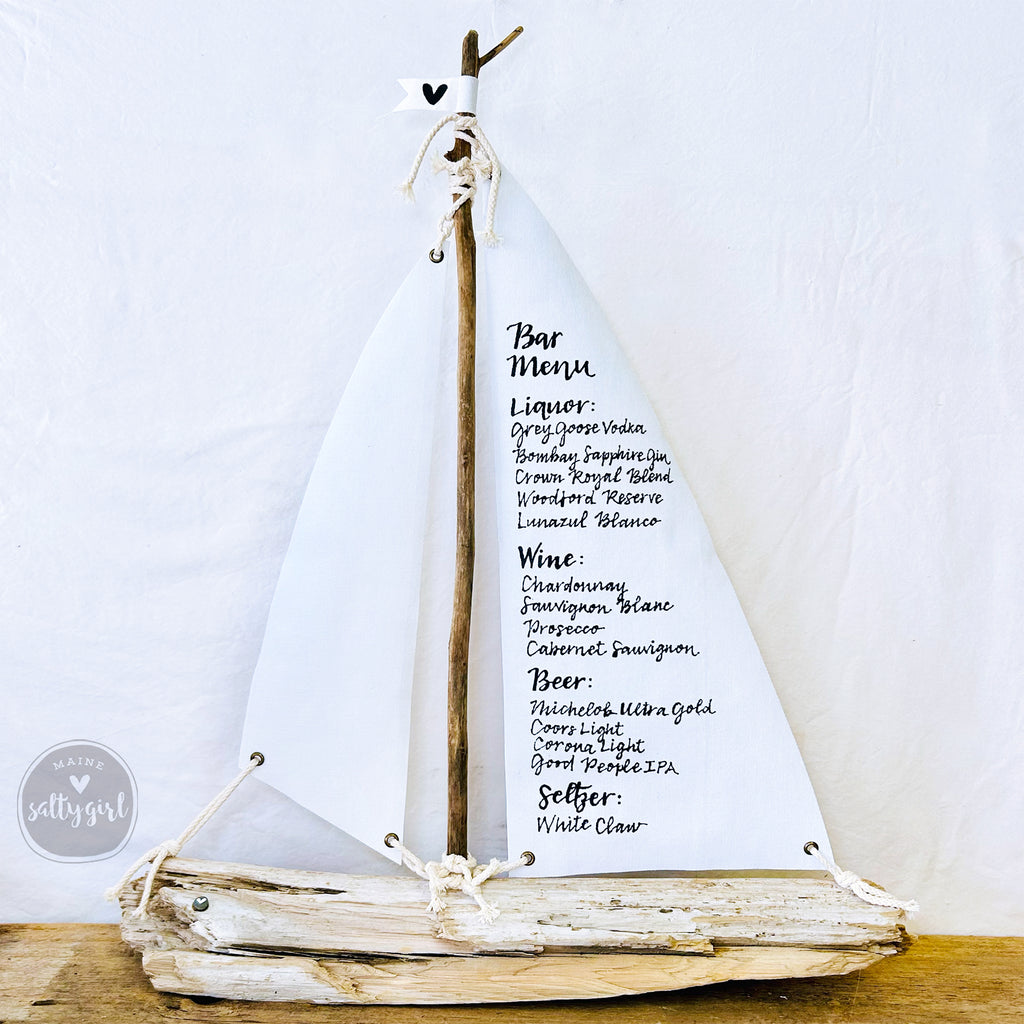 A white sailboat-shaped sign displaying a bar menu with various alcoholic and non-alcoholic drink options.