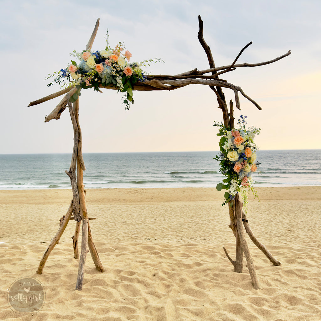A rustic wooden arch adorned with vibrant flowers stands on a sandy beach, with the ocean visible in the background.