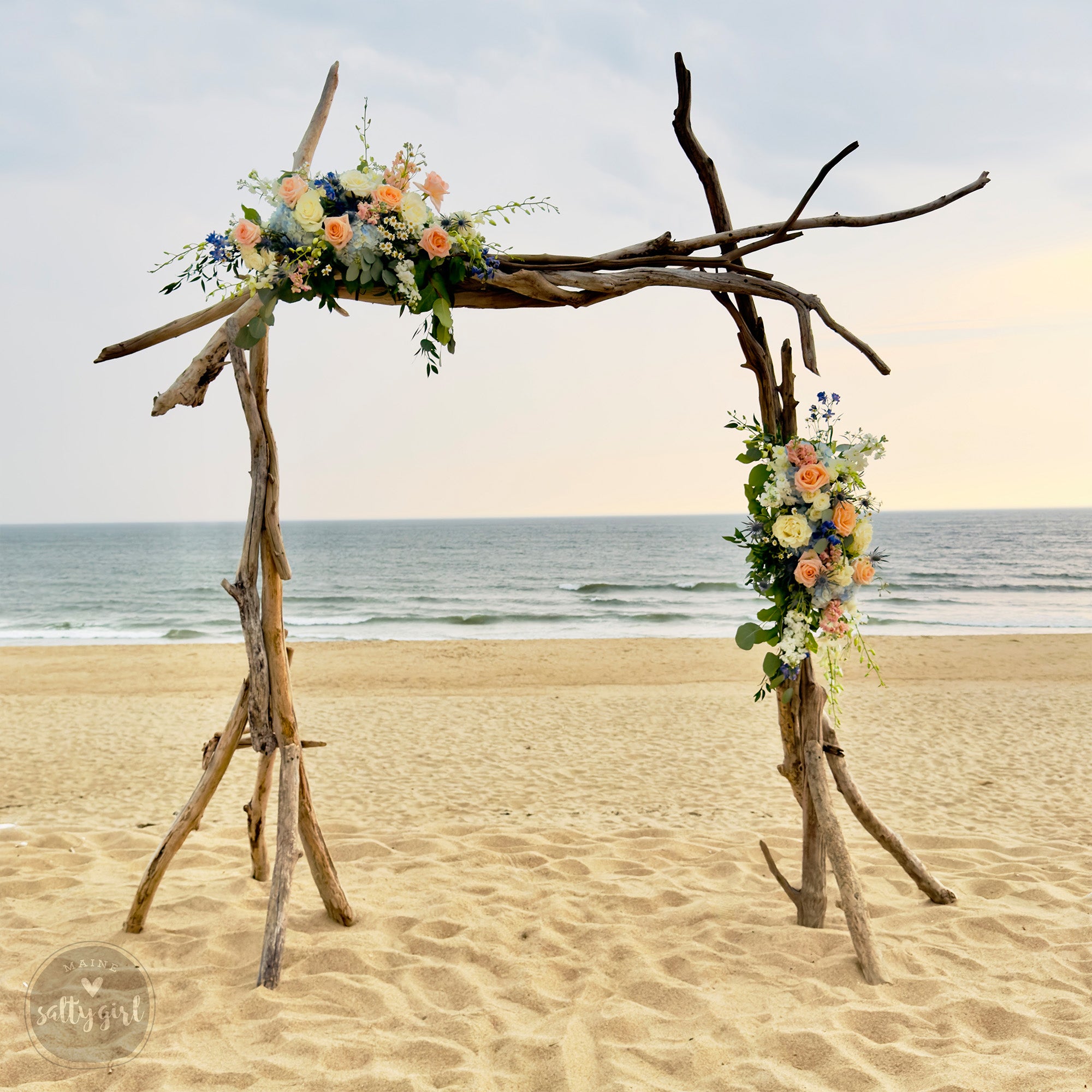 A rustic wooden arch adorned with vibrant flowers stands on a sandy beach, with the ocean visible in the background.