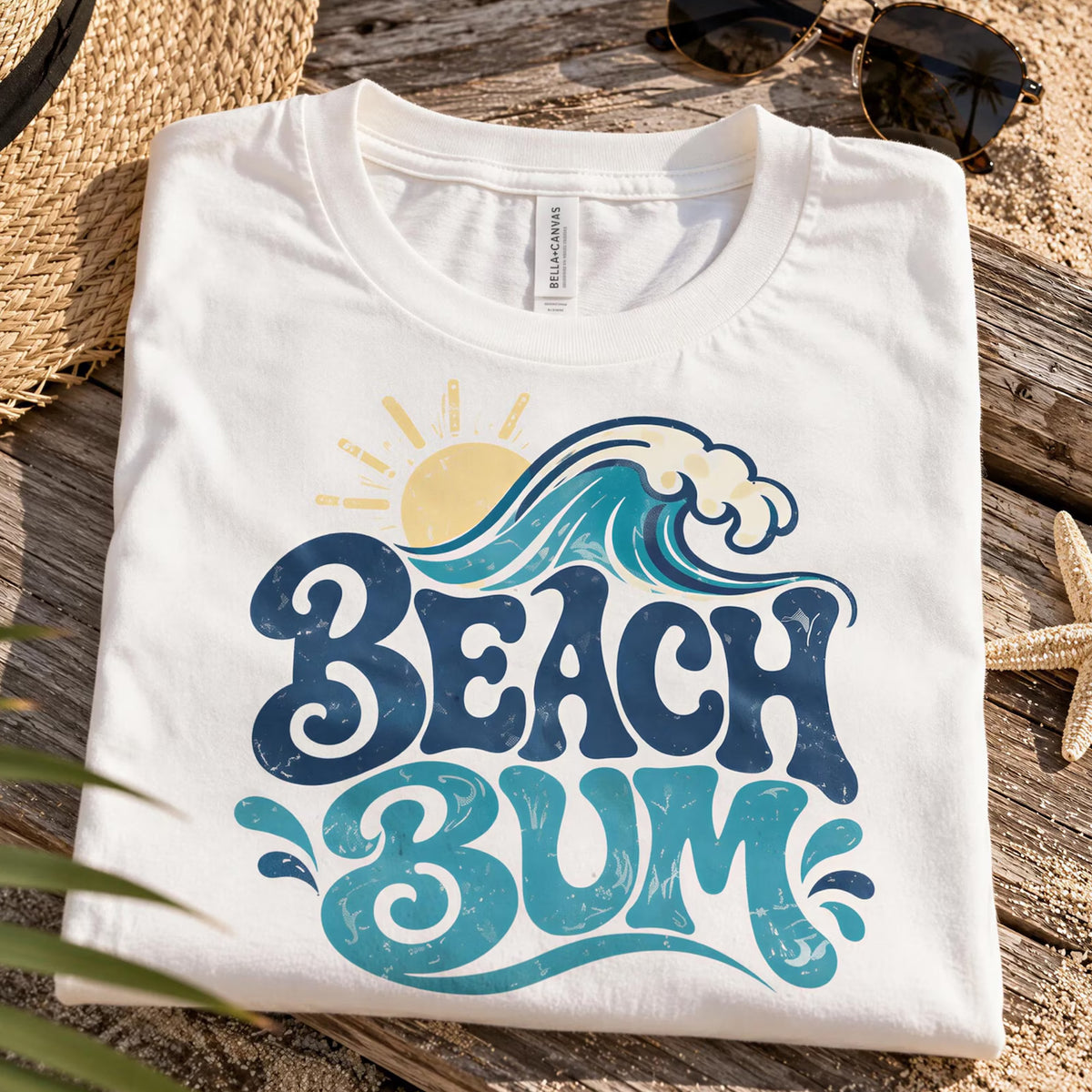 A white t-shirt with a graphic design of a wave and the text "BEACH BUM" printed on it, placed on a wooden surface with sunglasses and a straw hat nearby.