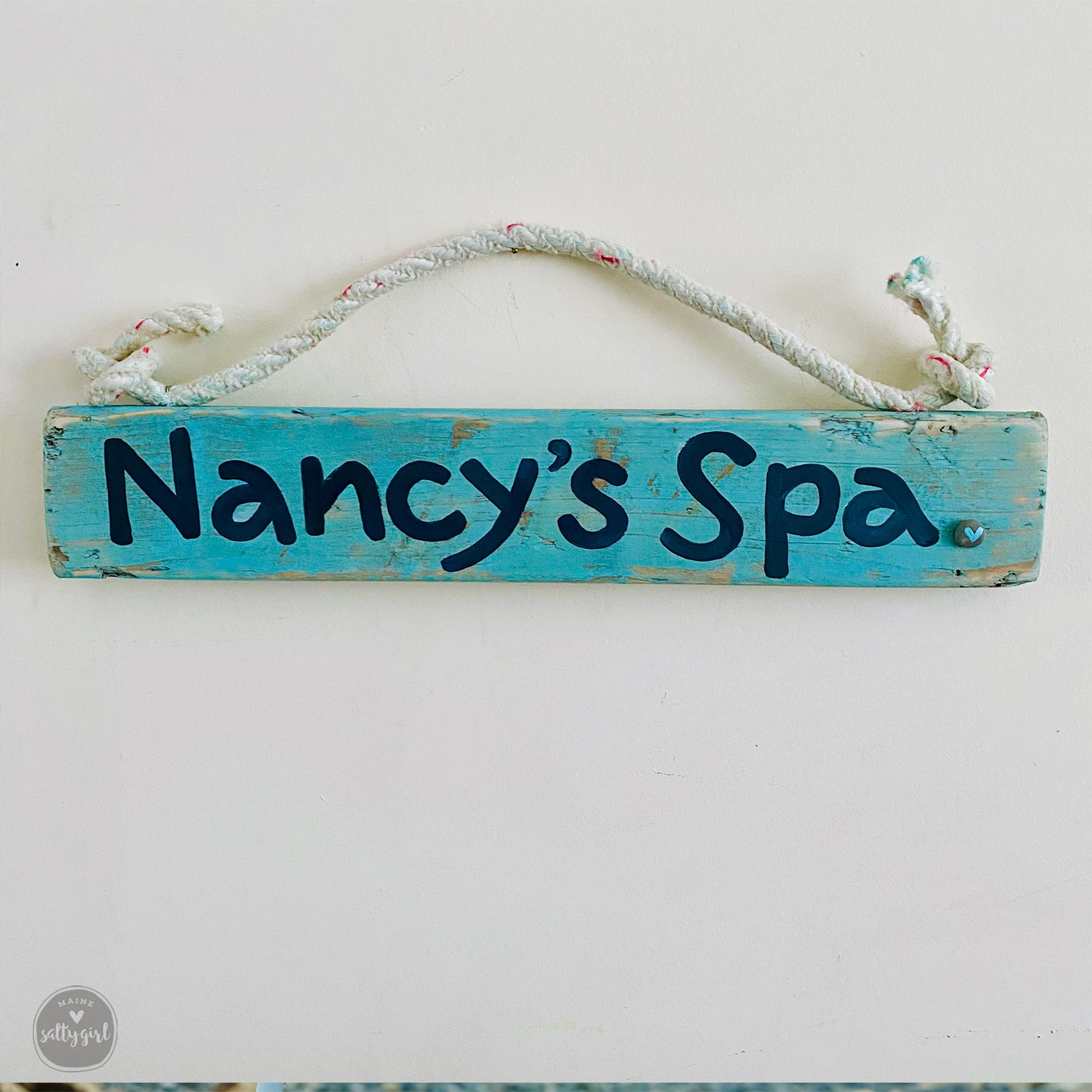 A blue wooden sign with the text "Nancy's Spa" hanging on a white wall.