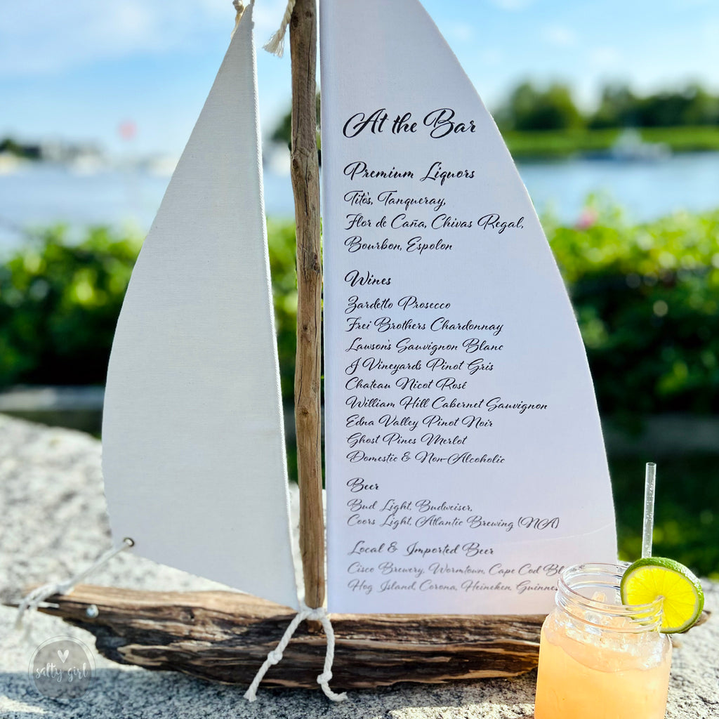 a menu board with a list of drinks and a glass of lemonade on a table, with a sailboat-shaped object in the foreground.