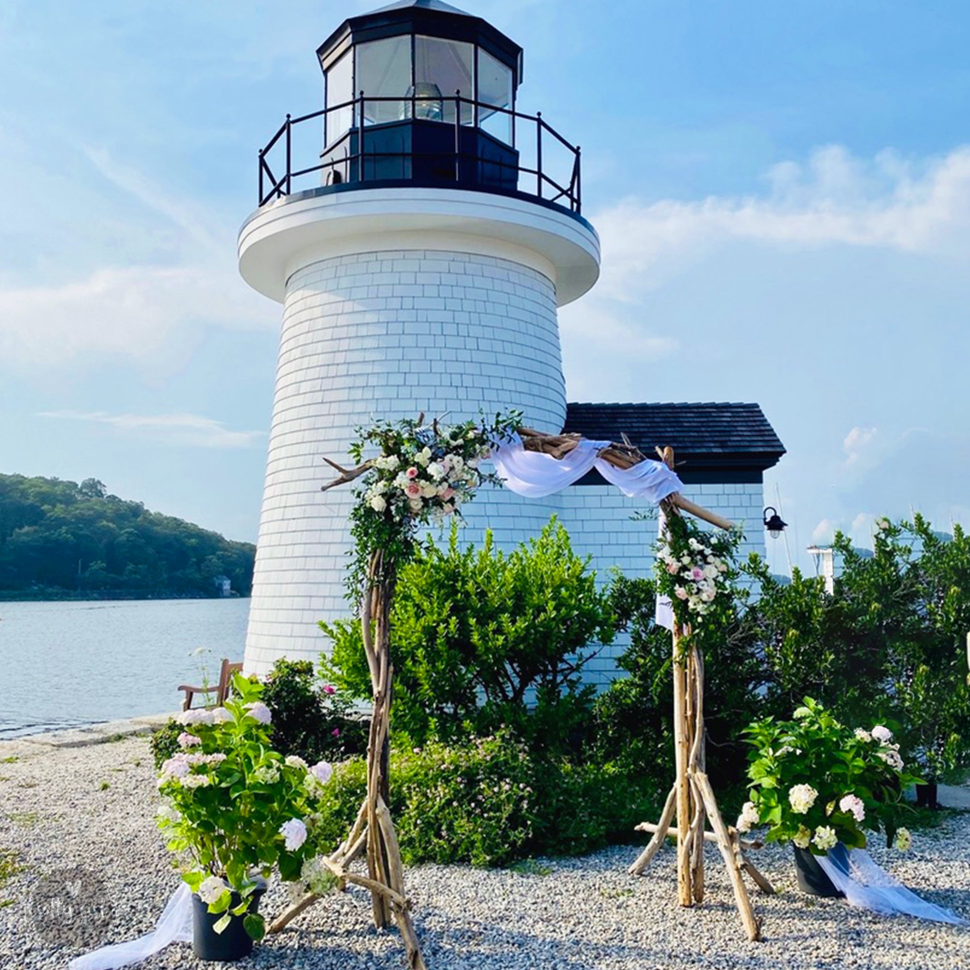 A white lighthouse with a black top and a small balcony, surrounded by lush greenery and flowers, with a wedding arch set up in front of it.
