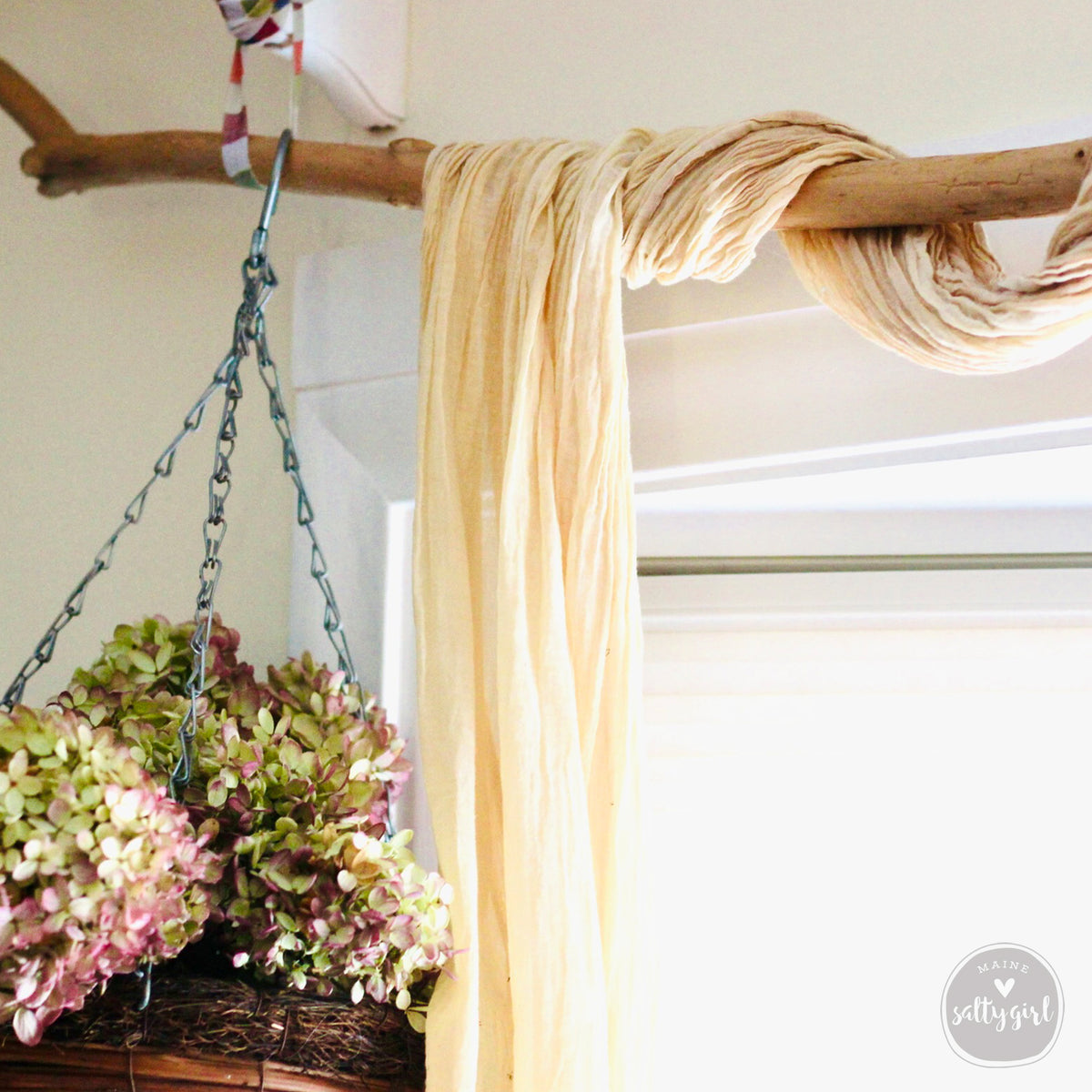 A hanging basket filled with green and pink flowers is suspended from a wooden branch, with a long yellow fabric draped over it.