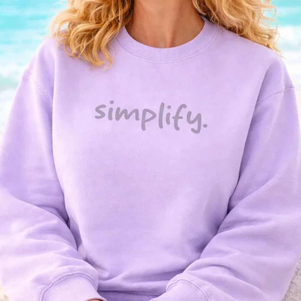 A smiling woman with curly blonde hair wearing a purple sweatshirt with the word "simplify" on it, sitting on a beach with the ocean in the background.