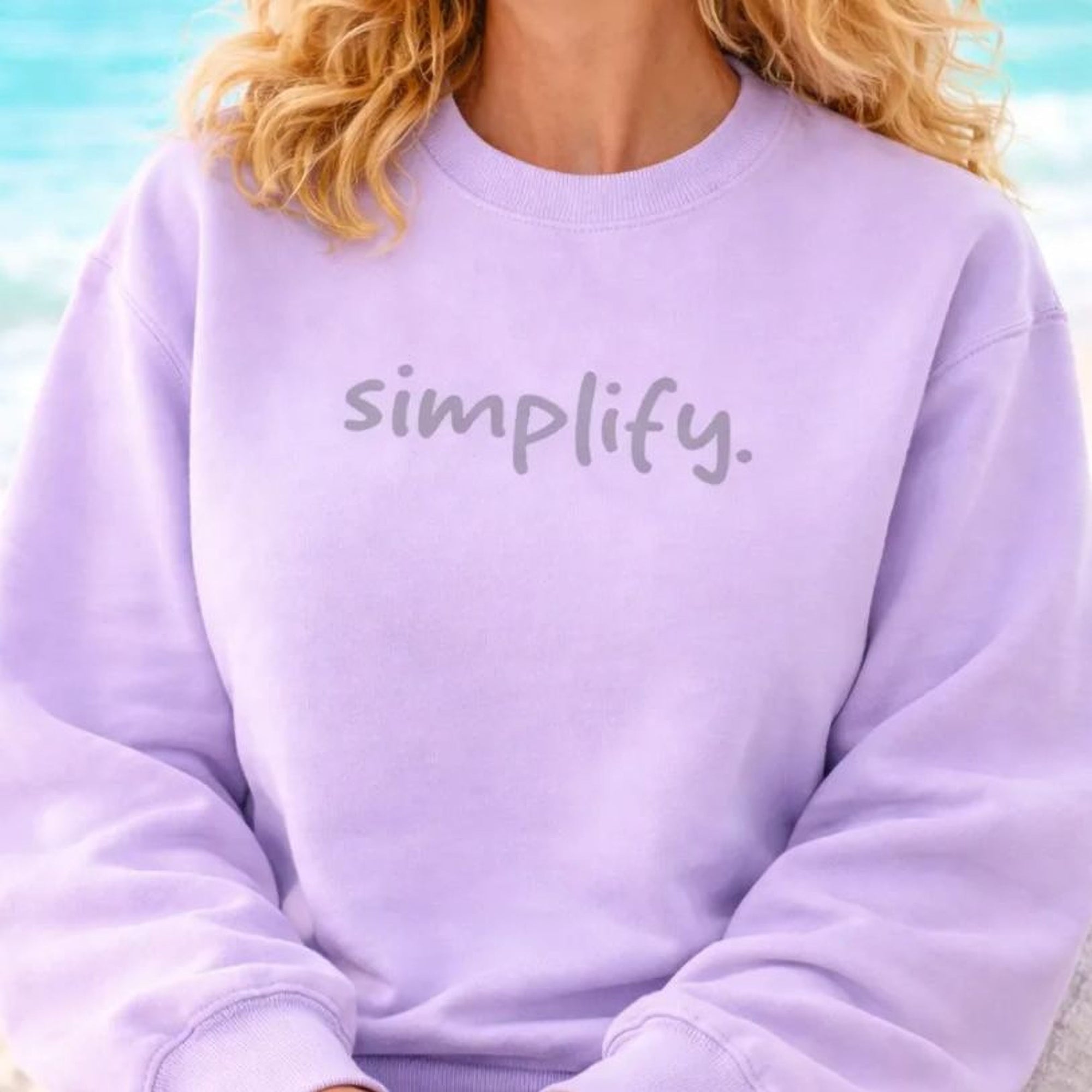 A smiling woman with curly blonde hair wearing a purple sweatshirt with the word "simplify" on it, sitting on a beach with the ocean in the background.