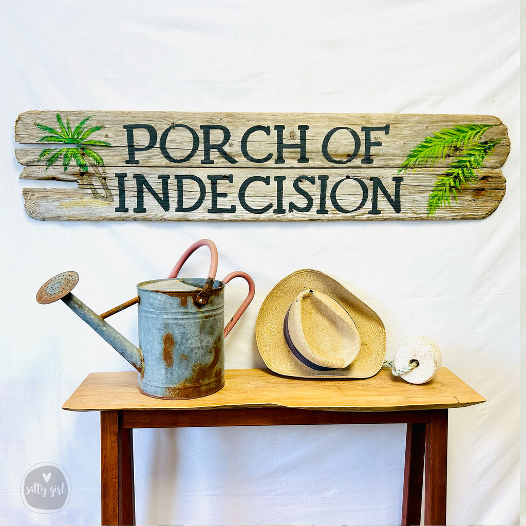 a rustic wooden sign with the text "PORCH OF INDECISION" and a watering can, along with a straw hat and a white ball, all displayed on a wooden table.