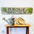 a rustic wooden sign with the text "PORCH OF INDECISION" and a watering can, along with a straw hat and a white ball, all displayed on a wooden table.