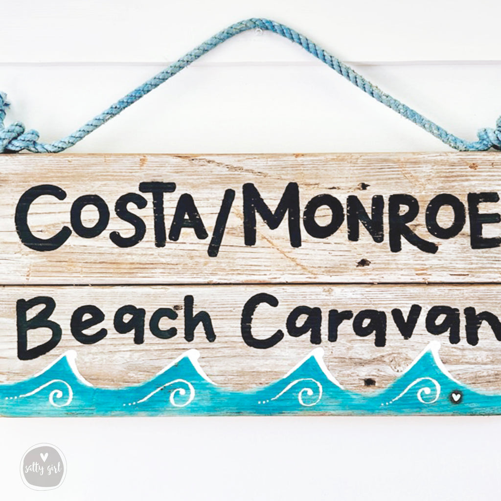 A wooden sign with the text "Costa/Monroe Beach Caravan" written on it, hanging on a wall with a blue rope.