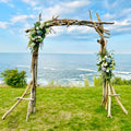 A rustic wooden archway adorned with flowers stands on a grassy field overlooking the ocean, with the blue sky and clouds in the background.