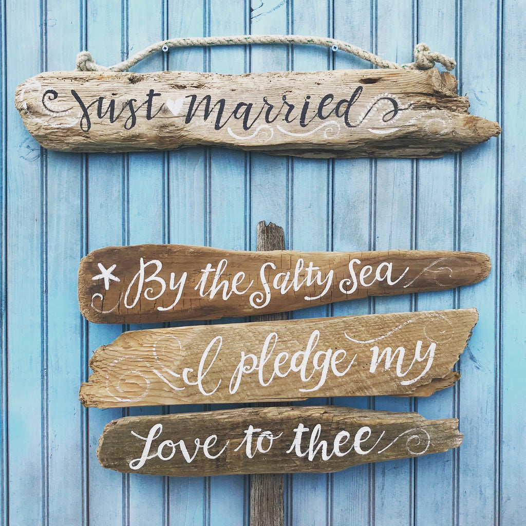 a blue wooden wall with a collection of driftwood signs. The signs are arranged in a row and display various phrases and sayings, including "Just Married", "By the Salty Sea", "Ipledge my love to thee", and "Love to thee".