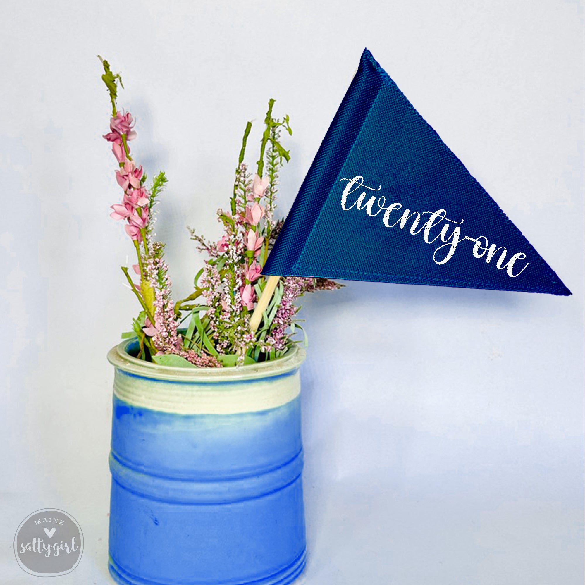 A blue ceramic vase filled with pink flowers and a blue triangular table number flag with the word "twenty-one" printed on it.