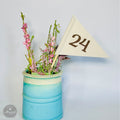 A blue vase filled with pink flowers and a white triangular table number flag with the number 24 on it, placed on a white surface.
