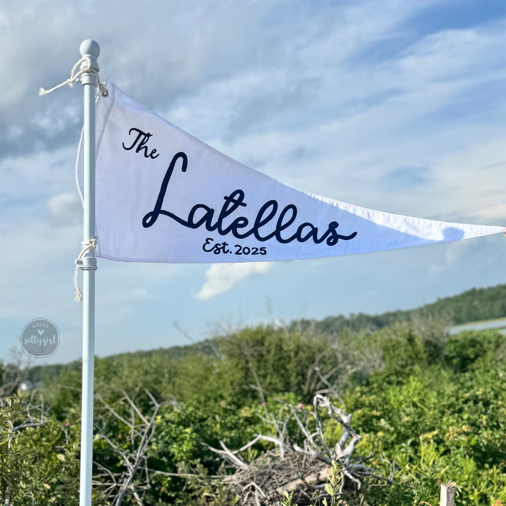 White flag with 'The Latellas est. 2025' text against a blue sky and green landscape.