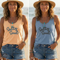 A woman wearing a peach Maine Salty Girl tank top with the text "Salty Soul" printed on it and denim shorts, standing on a beach with a hat on her head. Next to that is a woman wearing a denim blue Maine Salty Girl tank top with the text "Salty Soul" printed on it and denim shorts, standing on a beach with a hat on her head.