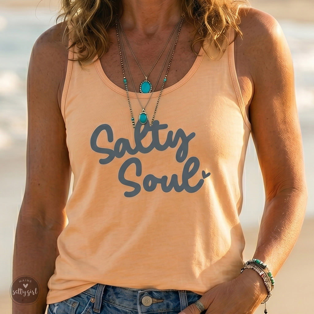 A woman wearing a peach Maine Salty Girl tank top with the text "Salty Soul" printed on it, standing on a beach.