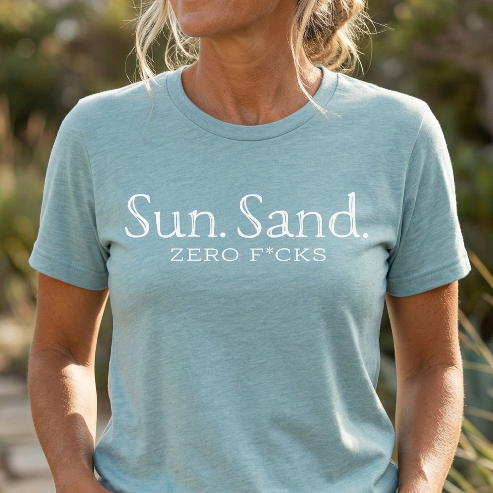 Person wearing a light blue t-shirt with 'Sun. Sand. ZERO F*CKS' text outdoors.