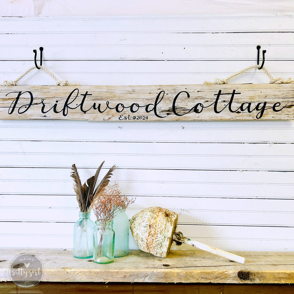 a rustic wooden sign that reads "Driftwood Cottage" with a vintage-style design, hanging on a white wall. Below the sign, there are three glass bottles with blue and green colors, and a small decorative item resembling a seashell.