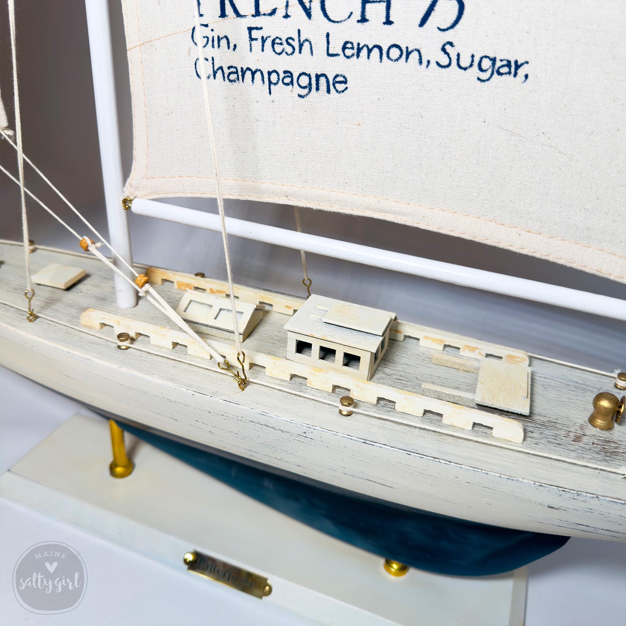A small white model ship with a banner that reads "French 7 Gin, Fresh Lemon, Sugar, Champagne" on its side.