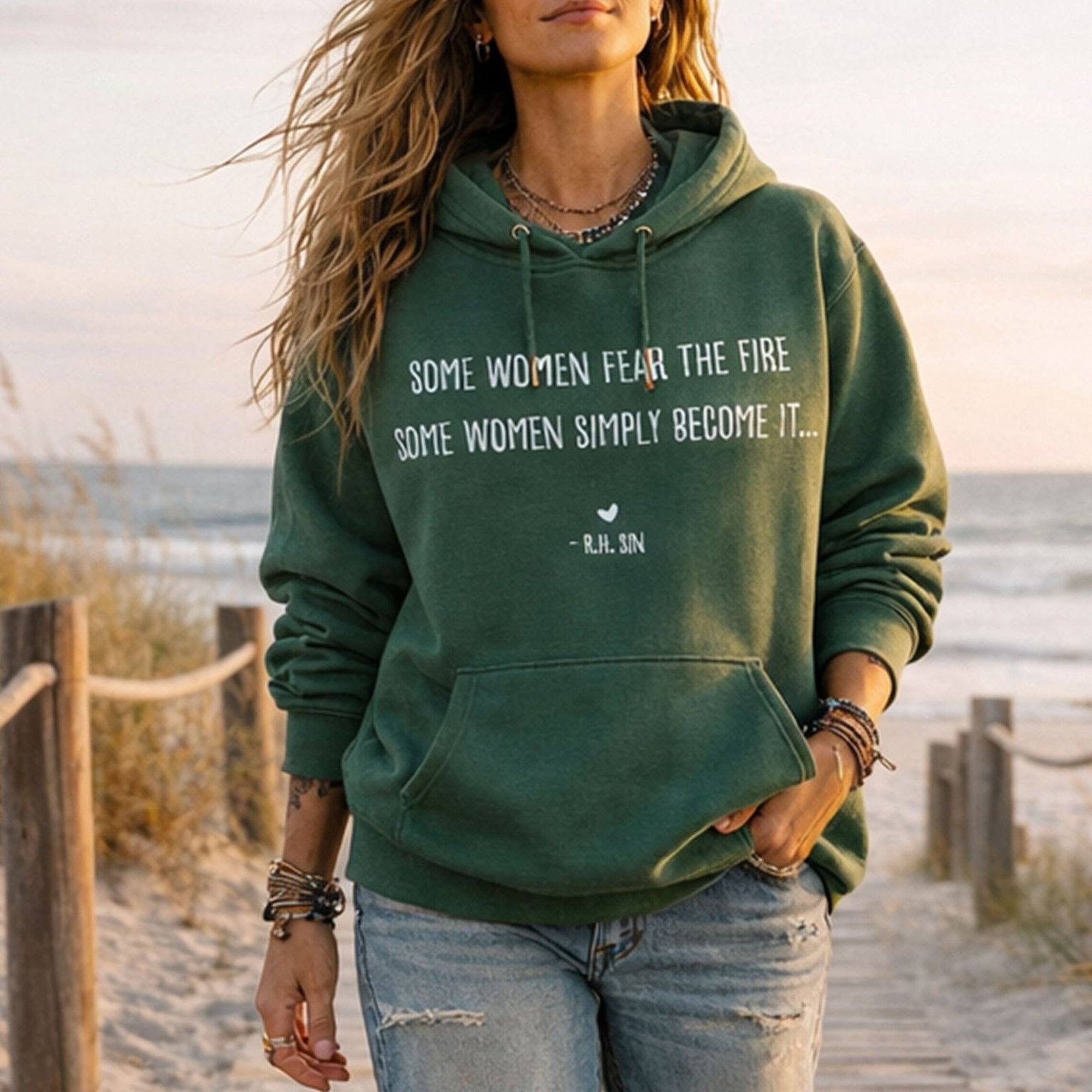 A woman wearing a green Maine Salty Girl hoodie with a quote printed on it, standing on a beach with the ocean in the background.