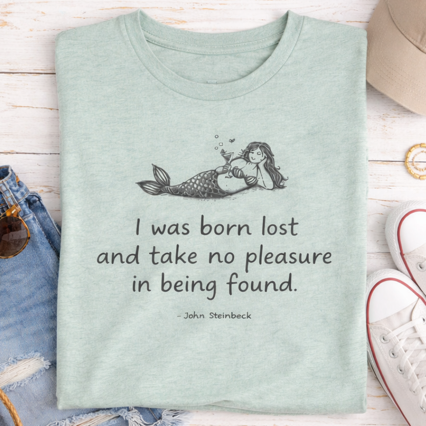 a green t-shirt with a mermaid graphic and a quote, a pair of jeans, a hat, sunglasses, and a pair of white sneakers, all laid out on a wooden surface.