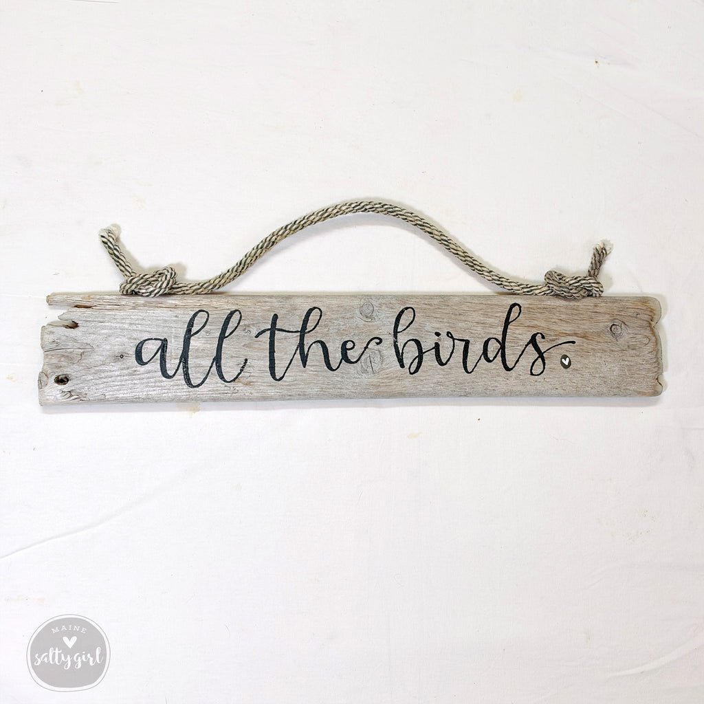A wooden sign with the text "all the birds" written on it, hanging from a rope.