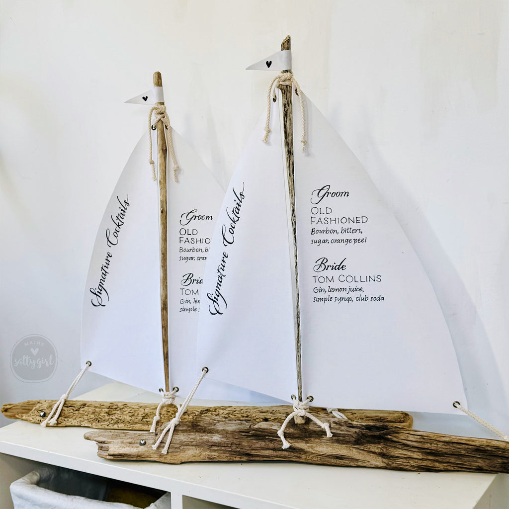 two white sailboat-shaped cocktail sails, each with a small flag attached to the top, displaying the names of cocktails and their ingredients.