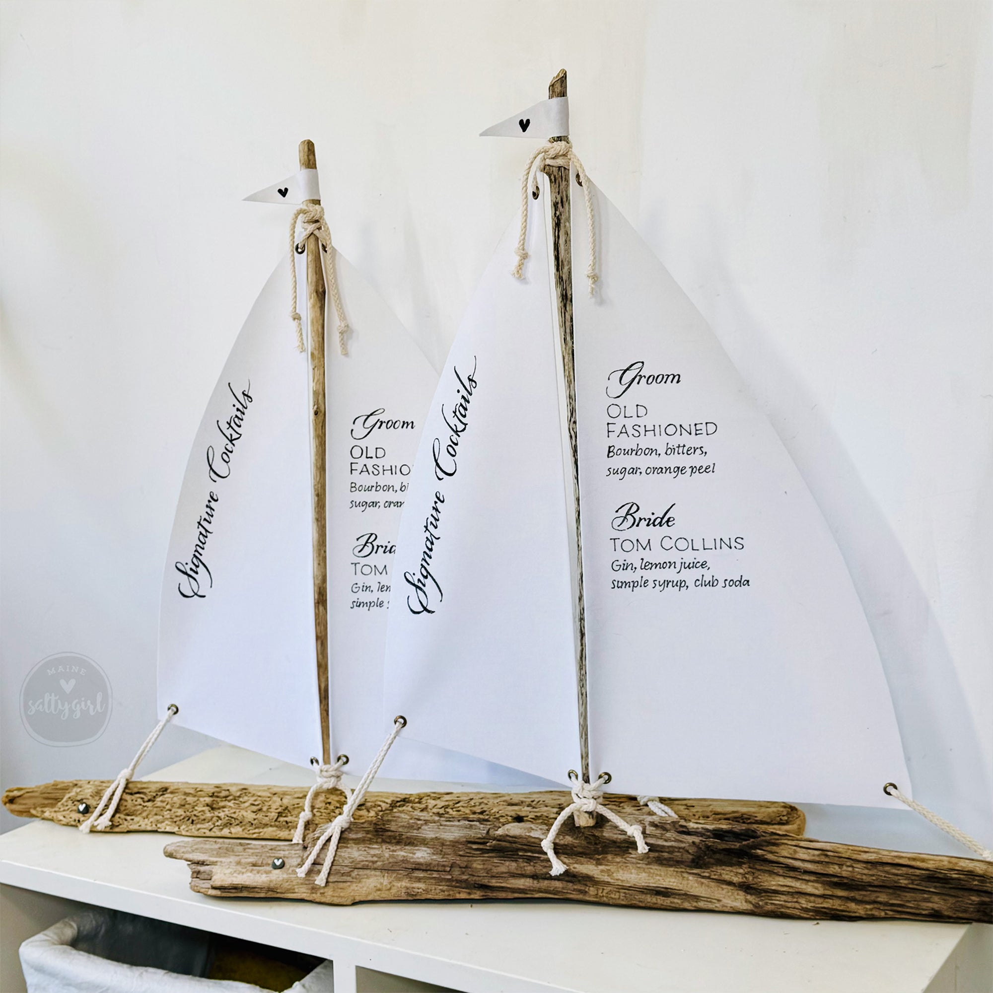 two white sailboat-shaped cocktail sails, each with a small flag attached to the top, displaying the names of cocktails and their ingredients.