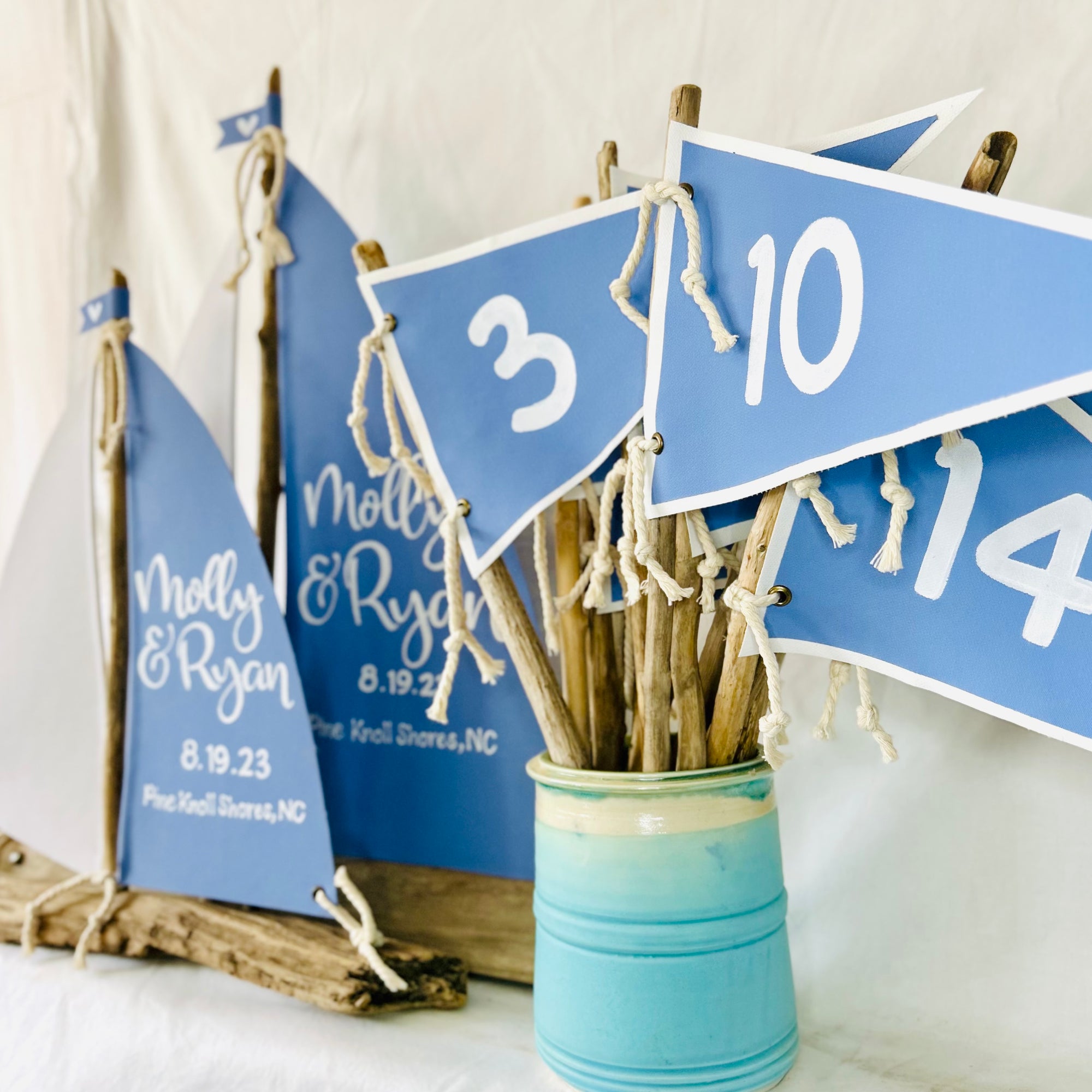 a blue sailboat with the names "Molly & Ryan" written on it, along with the date "8.19.23" and the location "Pine Knoll Shores, NC". The sailboat is placed next to a blue vase filled with wooden sticks, creating a nautical-themed display.