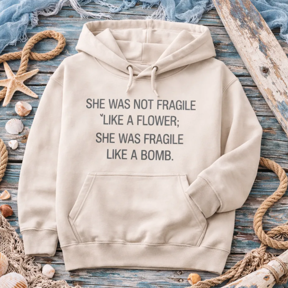 A beige hoodie with a humorous text printed on it, laid out on a wooden surface surrounded by seashells, rope, and starfish.