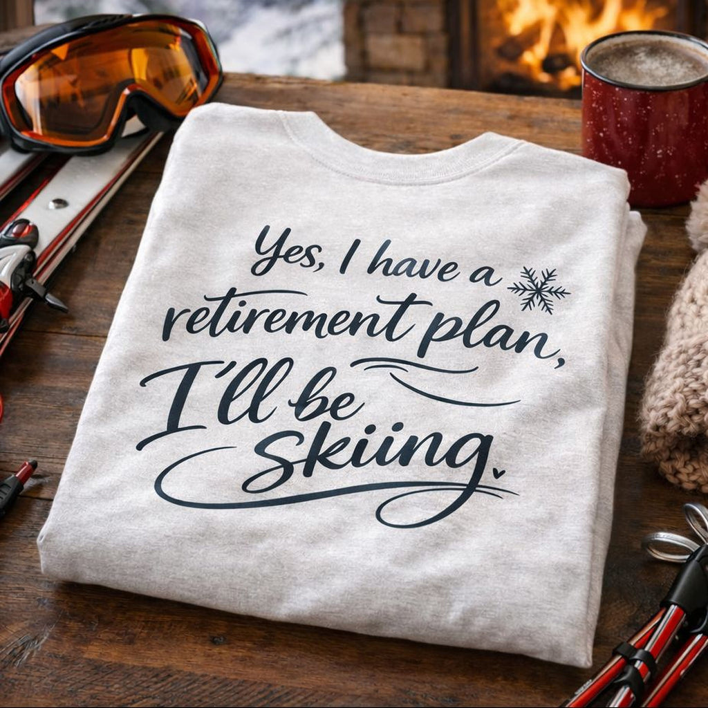 A white t-shirt with a humorous retirement plan and skiing message, along with ski gear and a mug, is displayed on a wooden surface.