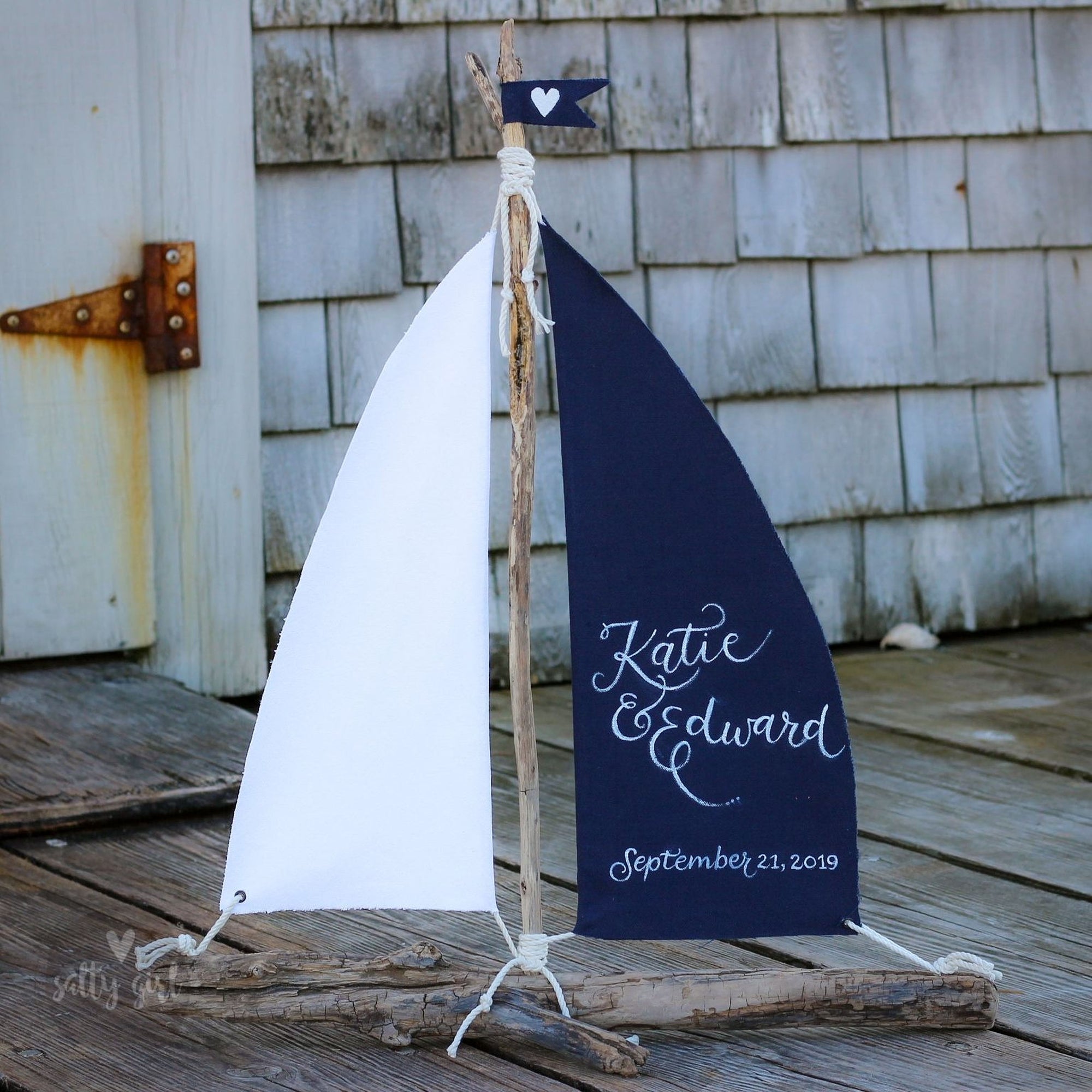 A white sailboat with a blue sail, featuring a small flag and a banner with the names 'Katie & Edward' and the date 'September 21, 2019'.