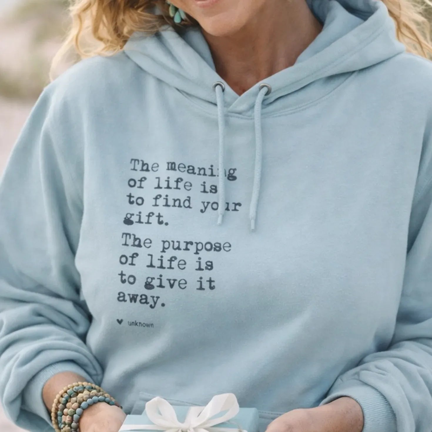 A smiling woman wearing a light blue hoodie with a quote printed on it, holding a gift box with a bow.