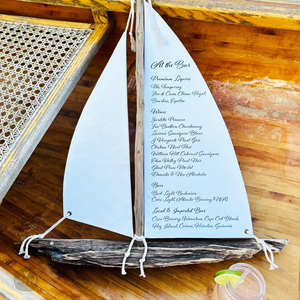 A white sailboat-shaped menu on a wooden table, with a glass of a beverage in the foreground.