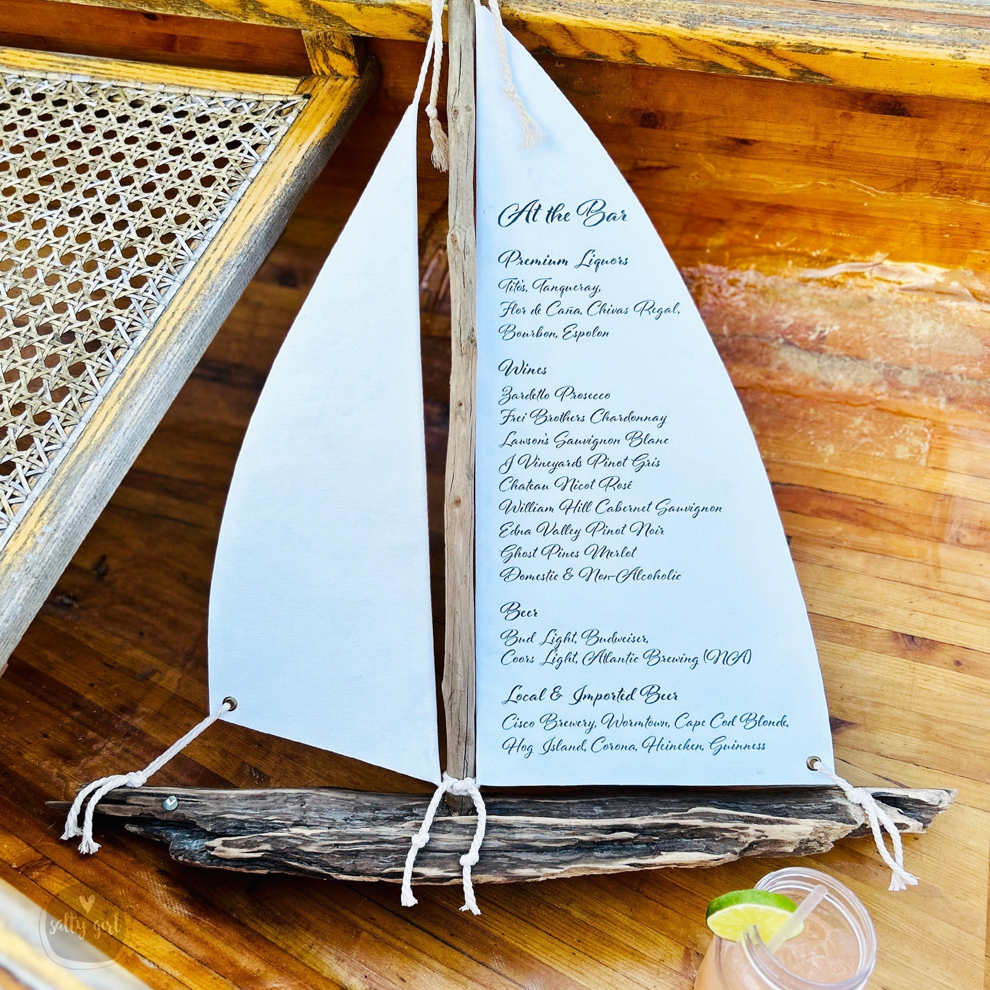 A white sailboat-shaped menu on a wooden table, with a glass of a beverage in the foreground.