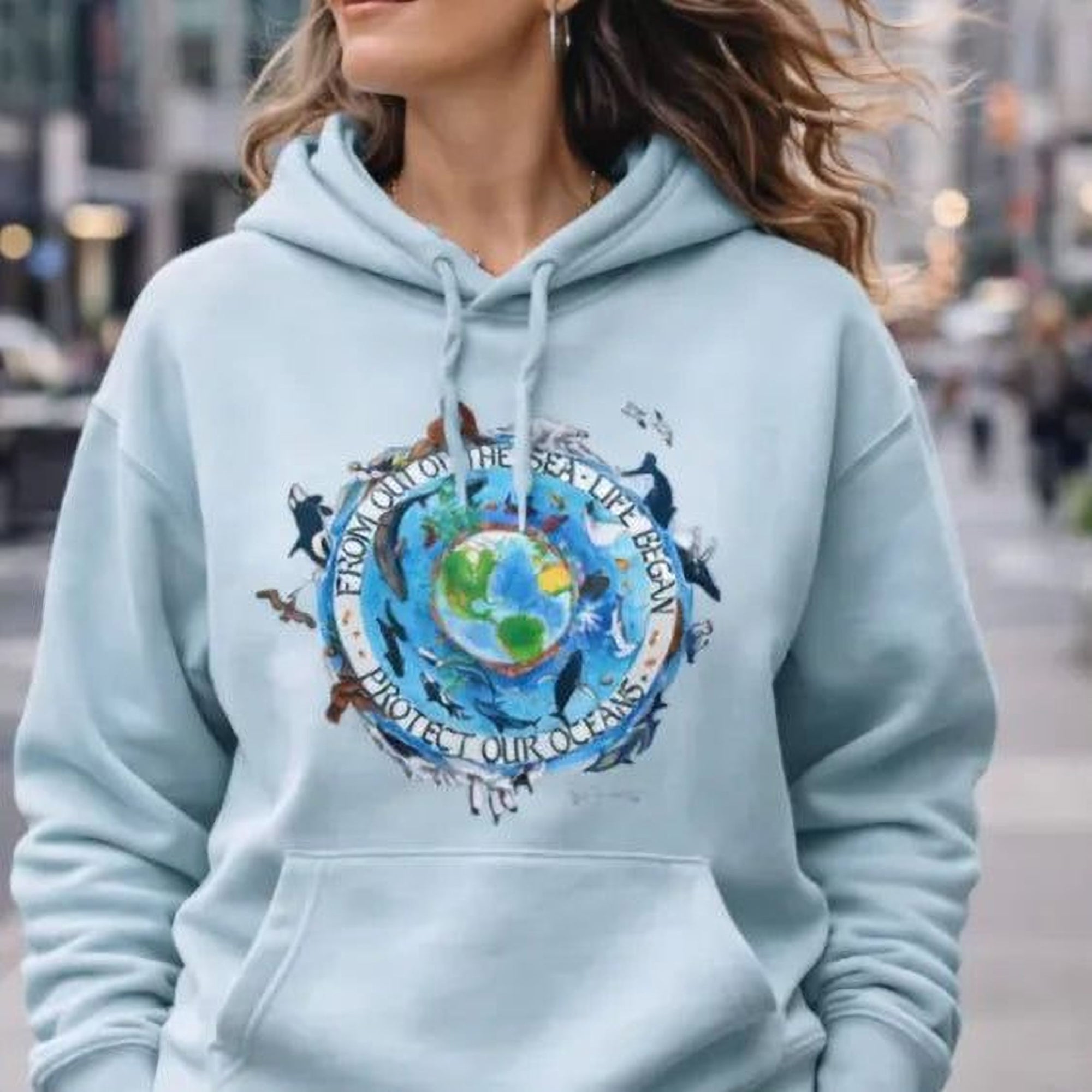 A woman wearing a light blue Maine Salty Girl hoodie with a graphic design of a globe and text, walking down a city street.