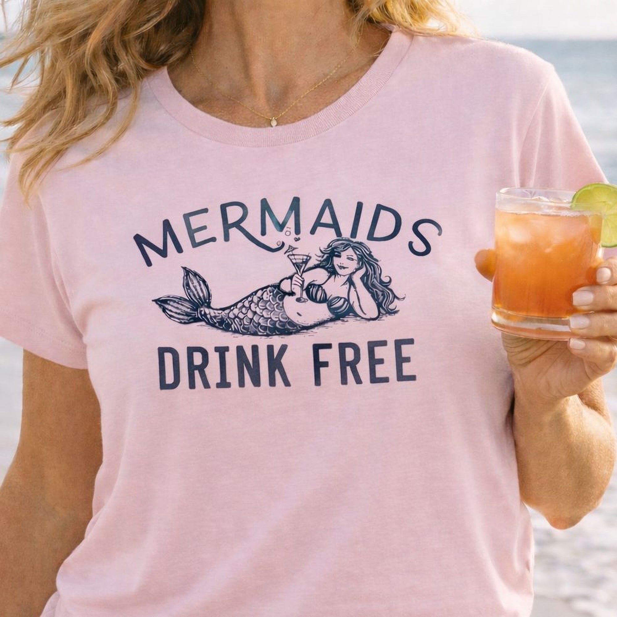 Person wearing a pink Maine Salty Girl t-shirt with 'Mermaids Drink Free' design, holding a drink on a beach.
