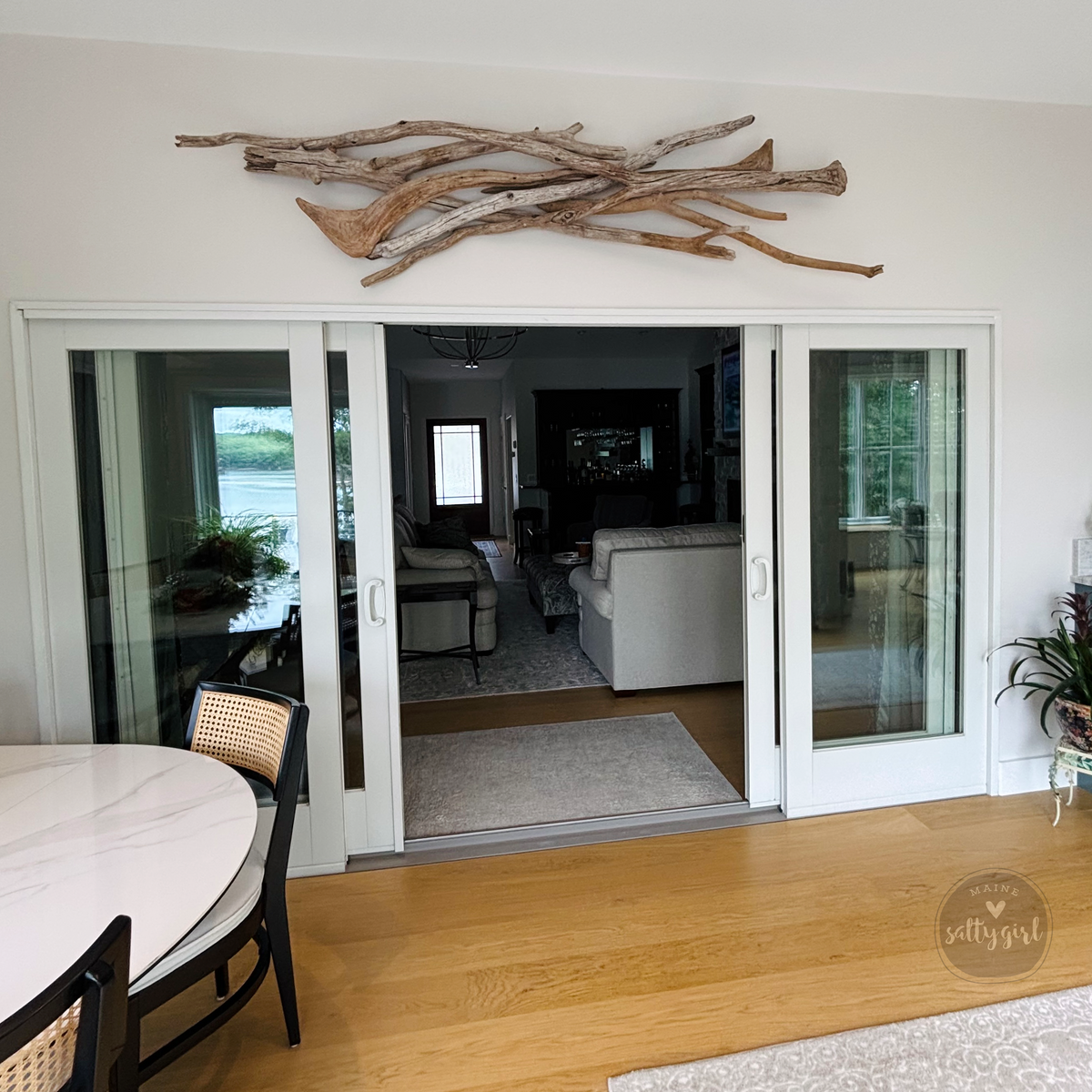 a modern living room with a large glass sliding door leading to another room. The sliding door is adorned with a wooden branch sculpture, adding a rustic touch to the space. The room features a white sofa, a coffee table, and a dining table with chairs. The overall design and decor create a harmonious and inviting atmosphere.