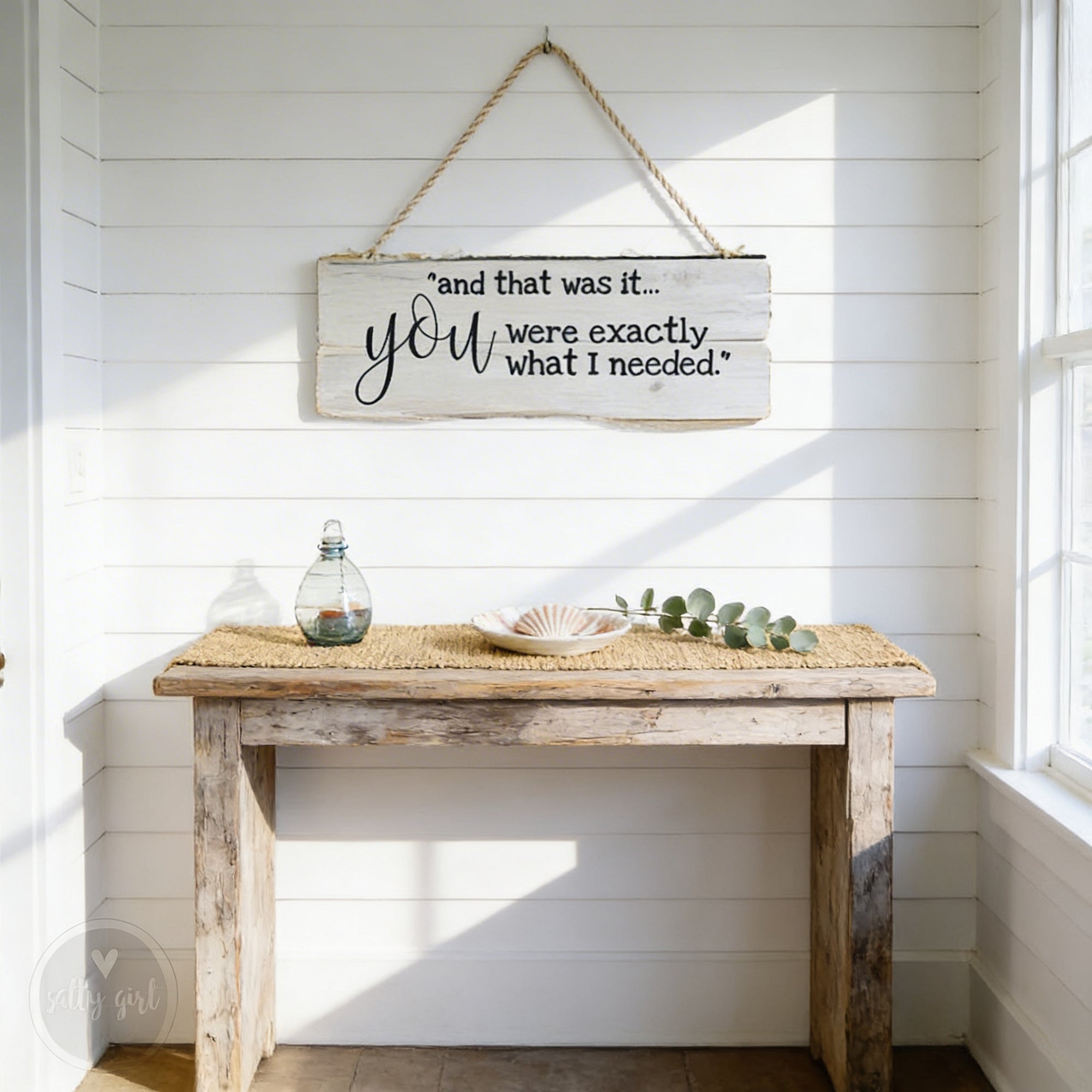 a rustic wooden table with a woven placemat, a glass bottle, a seashell, and some greenery, all set against a white wall with a hanging sign.