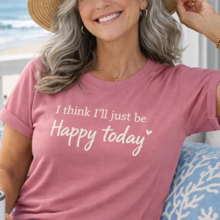 A smiling woman wearing a straw hat and a pink t-shirt with the text "I think I'll just be Happy today" is sitting on a bench near the ocean.