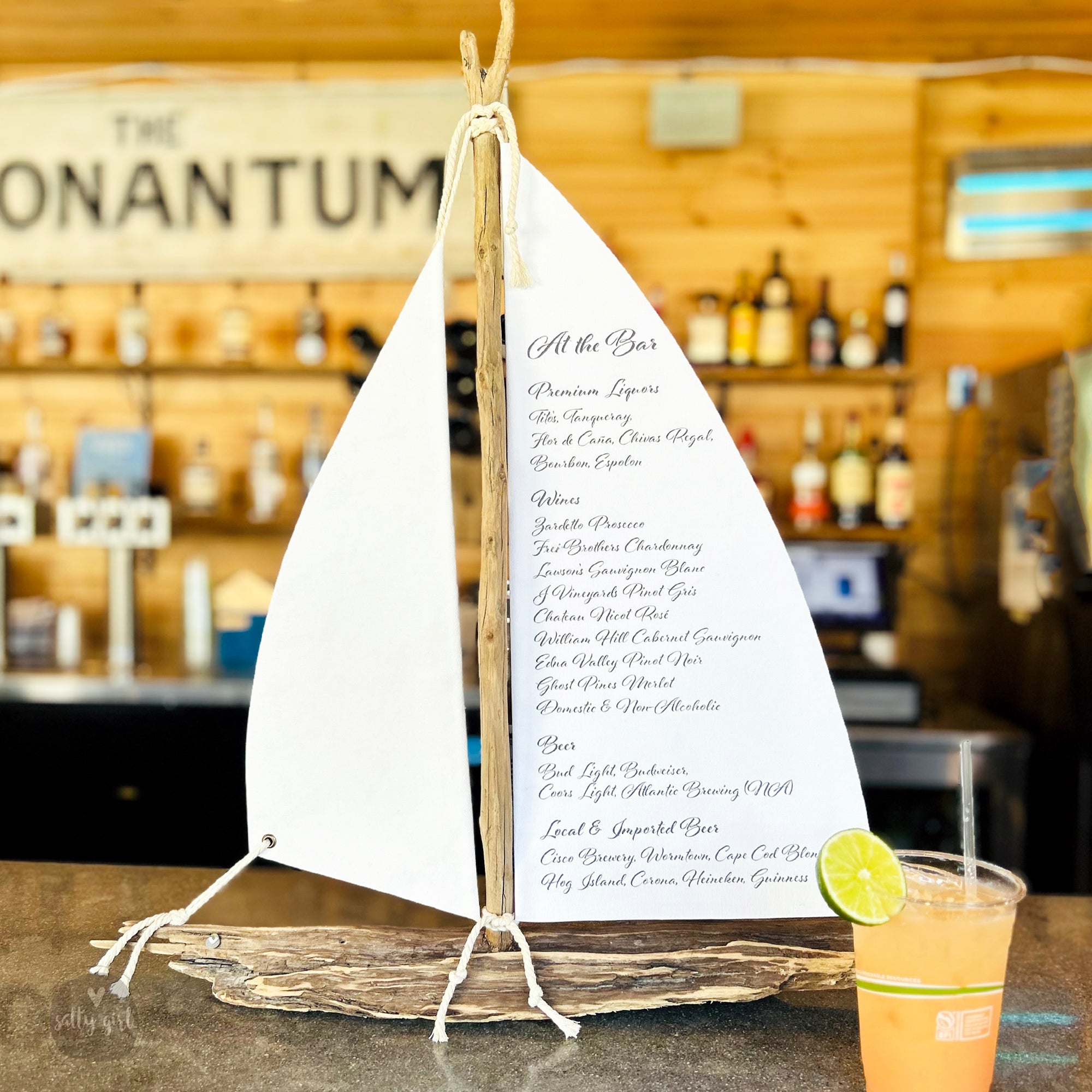 A wooden sailboat-shaped object with a list of drinks on it, placed on a bar counter next to a drink in a plastic cup.