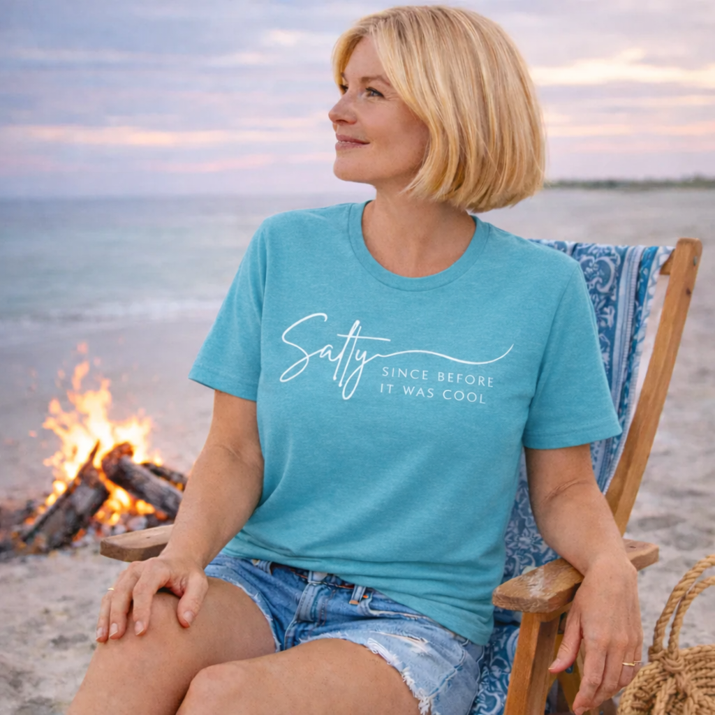 A woman in a blue t-shirt and denim shorts sits on a beach chair, smiling and looking towards the ocean.
