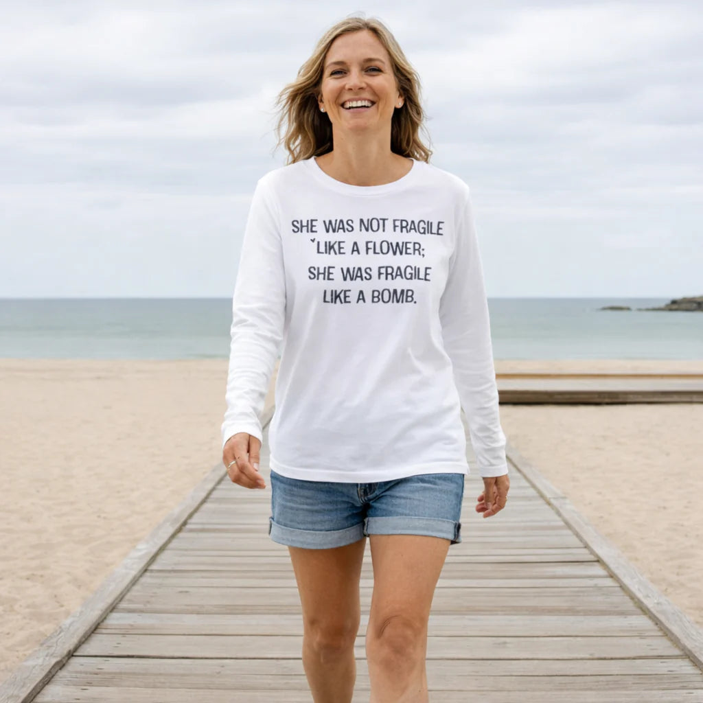 A woman wearing a white long-sleeved shirt with text printed on it, walking on a wooden boardwalk near the ocean.