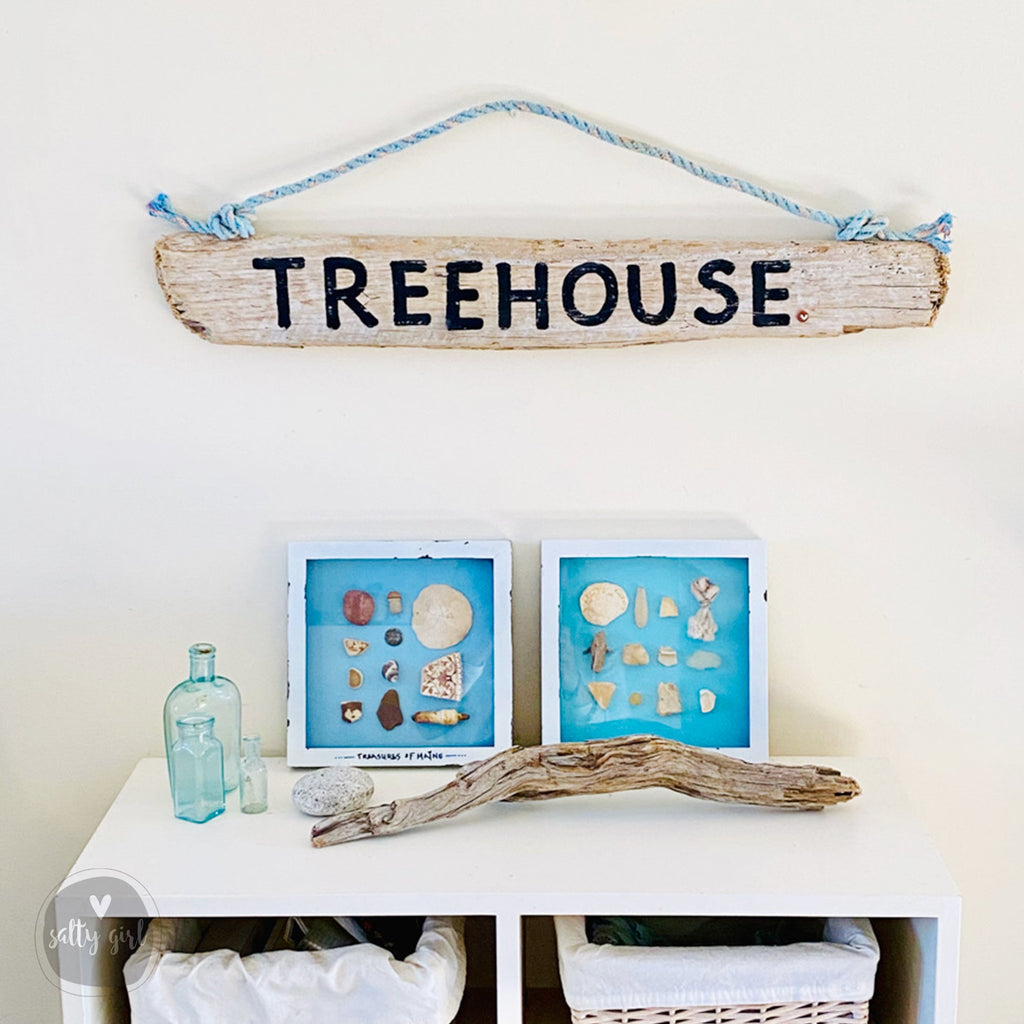 a rustic wooden sign that reads "TREEHOUSE" hanging on a wall, with two framed pictures of seashells placed below it. There is also a driftwood branch and a blue bottle on a white shelf below the framed pictures.
