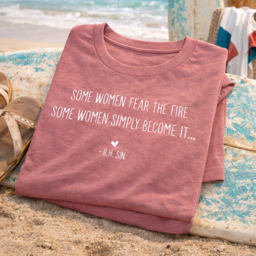 A pink t-shirt with a quote about women and fire is displayed on a beach, with a pair of sandals and a towel visible in the background.
