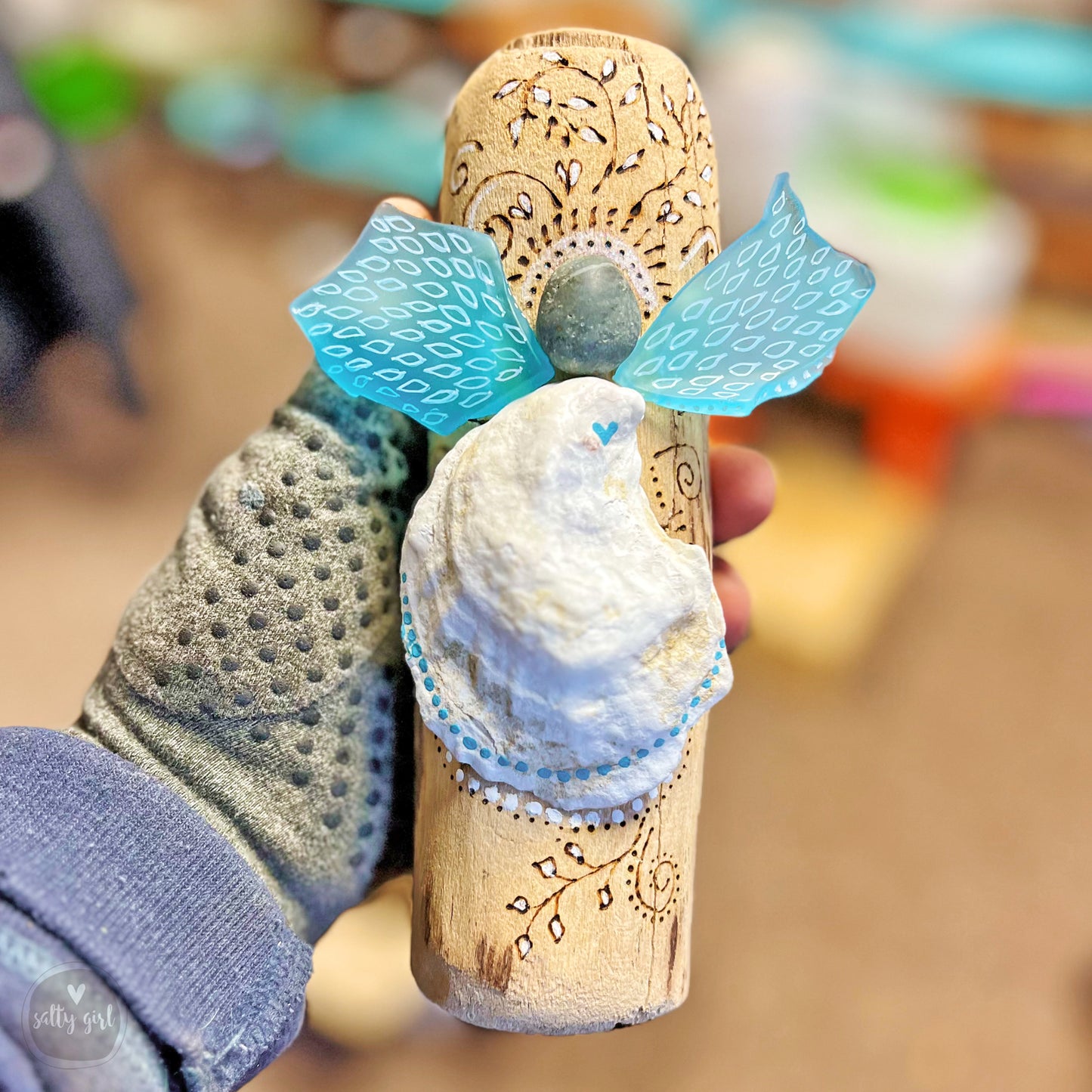 A hand holding a wooden cork with a blue and white design, including a small white bird-like figure and a stone or gemstone.