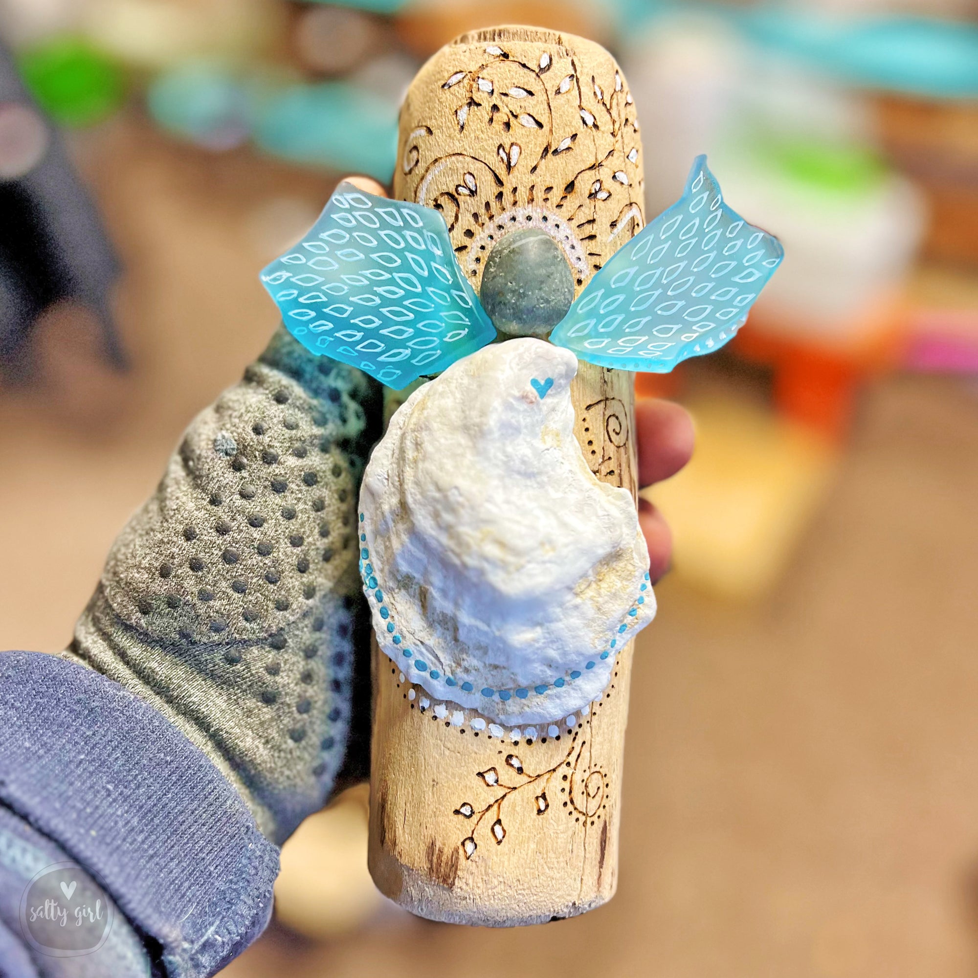 A hand holding a wooden cork with a blue and white design, including a small white bird-like figure and a stone or gemstone.