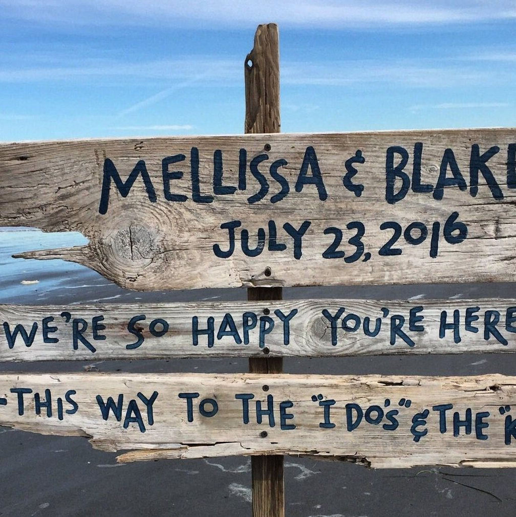 A wooden sign with the names "Mellissa & Blake" and the date "July 23, 2016" written on it, along with the phrase "We're so happy you're here" and "This way to the 'I do' & the 'I do'".