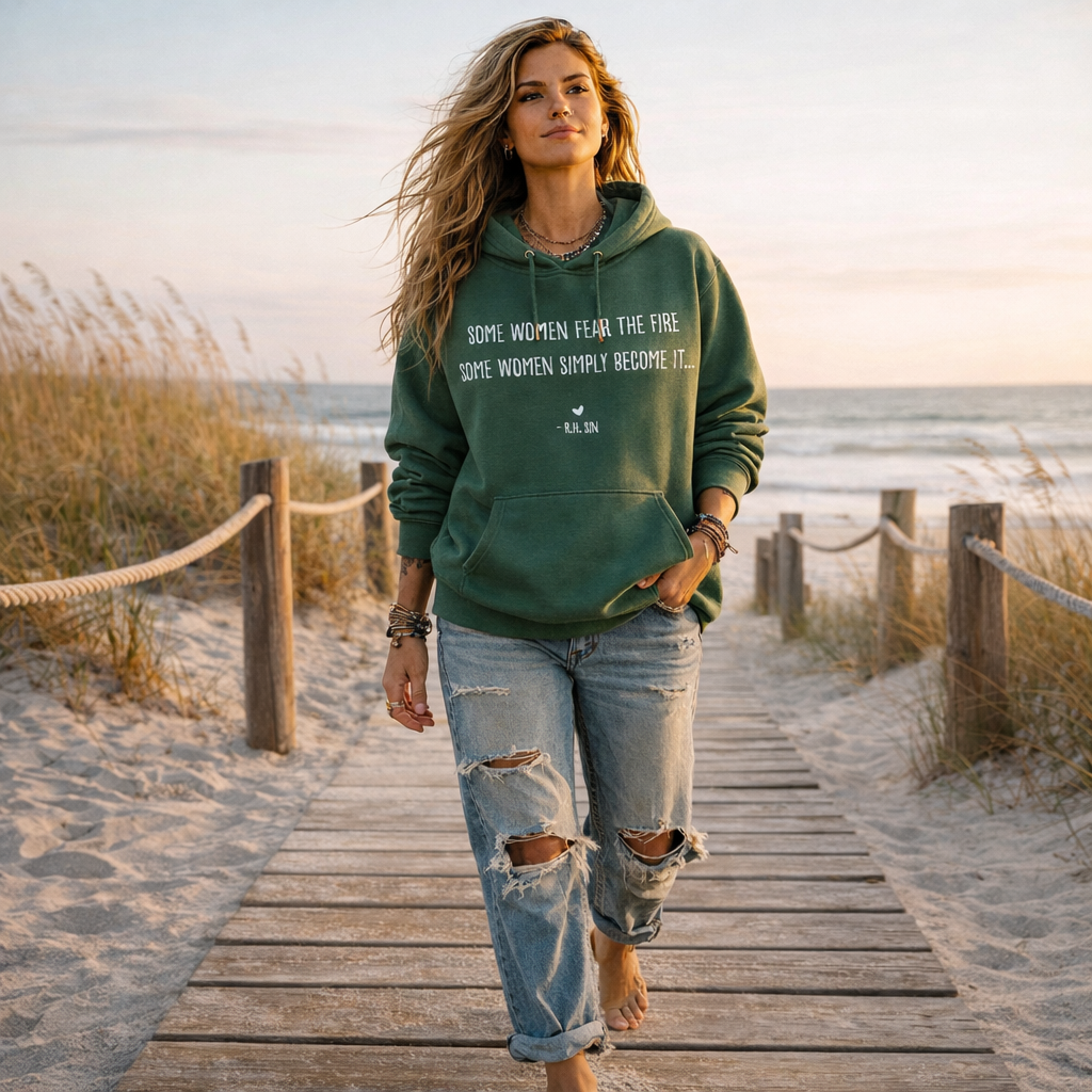 A woman wearing a green hoodie with text on it walks on a wooden boardwalk near the ocean.