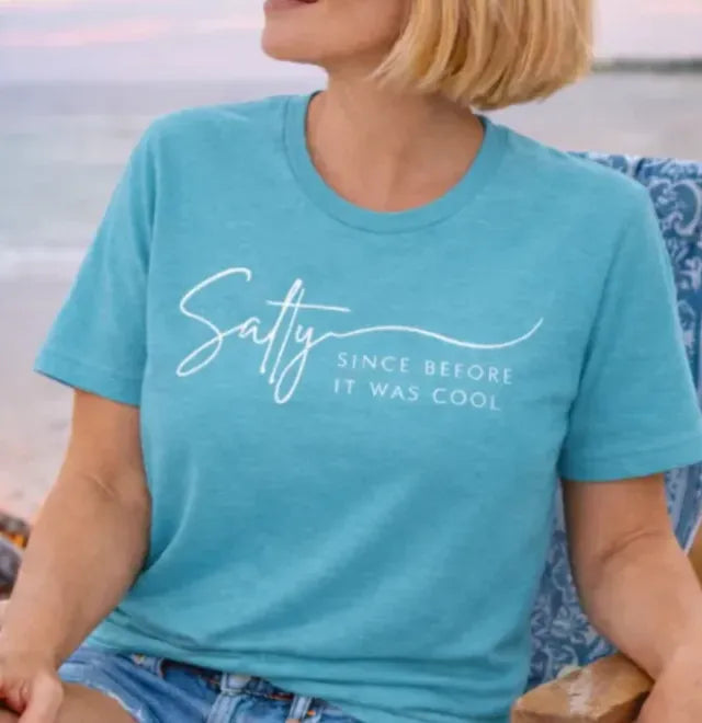 A woman in a blue t-shirt and denim shorts sits on a beach chair, smiling and looking towards the ocean.